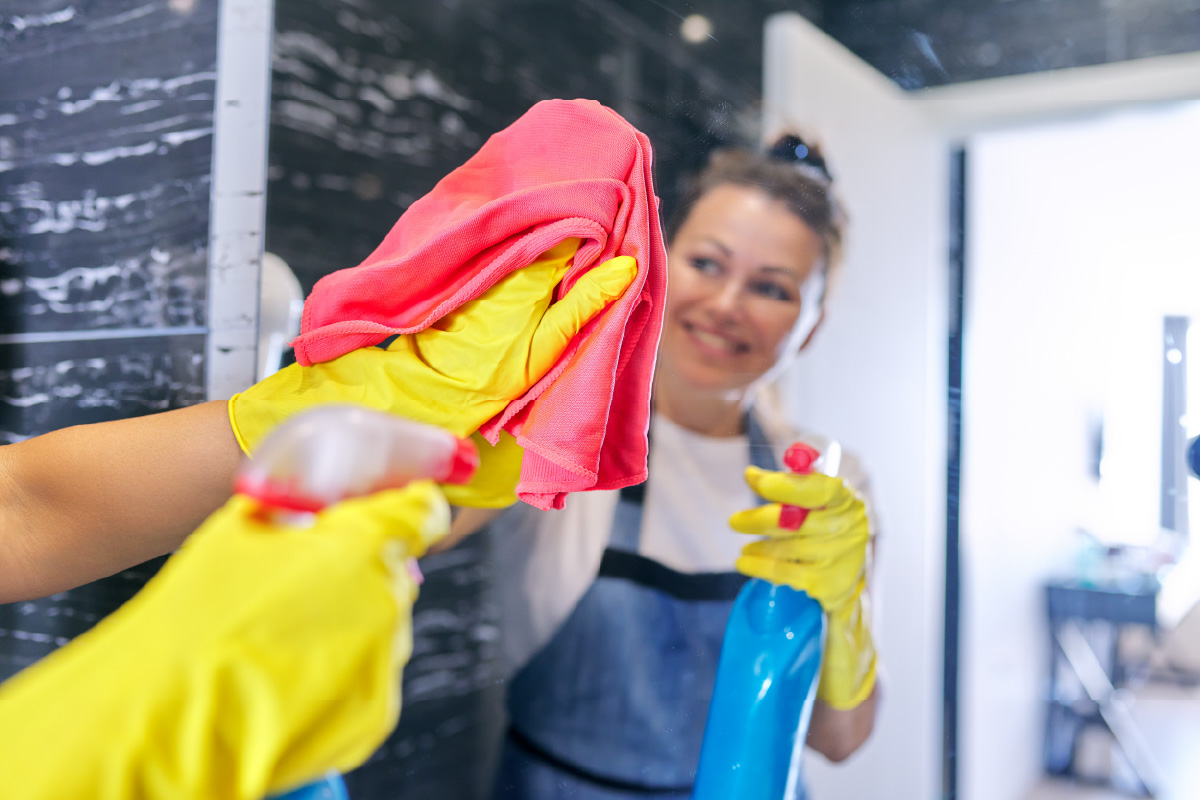 Reinigungskraft am Spiegel abwischen mit Glasreiniger und Mikrofasertuch in der Hand